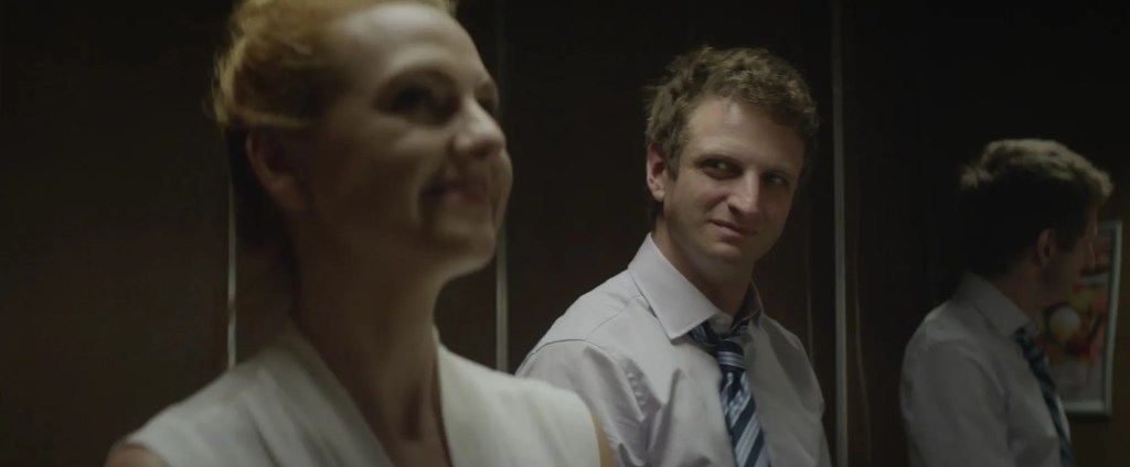 A still from Overtime portraying the male protagonist standing in an elevator with a female co-worker. She's out of focus, smiling and looking ahead while the man is looking at her with an ambiguous facial expression. 