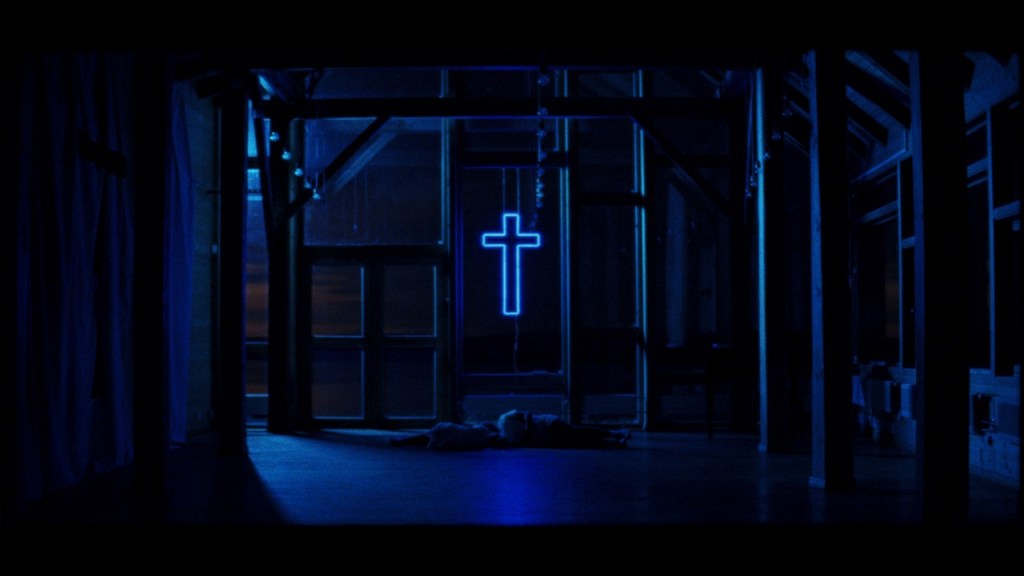 Still from Children of Satan showing a house with many windows during nighttime and a neon sign in the shape of a cross centered in the middle that shines a bluish light over its surroundings. 