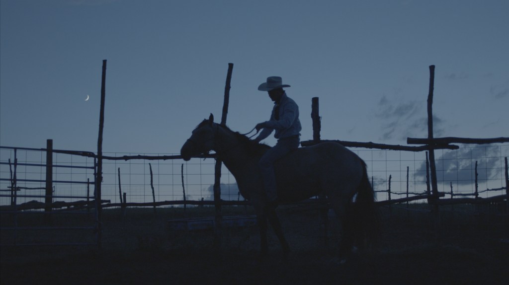 Brady (Brady Jandreau) from The Rider, wearing his signature cowboy hat, riding his horse late one evening, with fences, a dark bluish sky, and the crescent moon visible behind him.