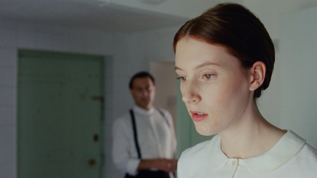 A still from The Sermon showing the female protagonist in focus looking ahead of herself with an empty facial expression. Out of focus behind her is a man standing, looking at her. 