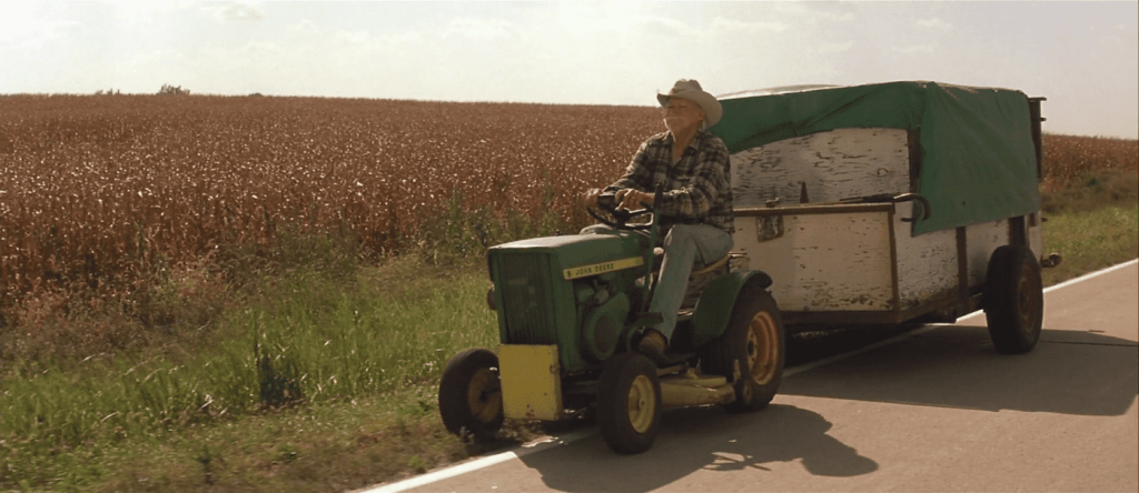 Alvin (Richard Farnsworth) from The Straight Story riding alongside the road on his green lawn mower and its accompanying trailer. He's smiling and wearing a beige cowboy hat, a checkered shirt, and light blue jeans. Beside him are fields of vegetation in earthy and warm hues of brown. The sun is shining.