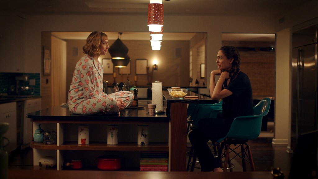 Still from Waffle showing the two female protagonists sitting in the kitchen, one of them on top of a kitchen counter and the other in front of her in a chair. They are, while both wearing pyjamas, looking at each other and smiling. 