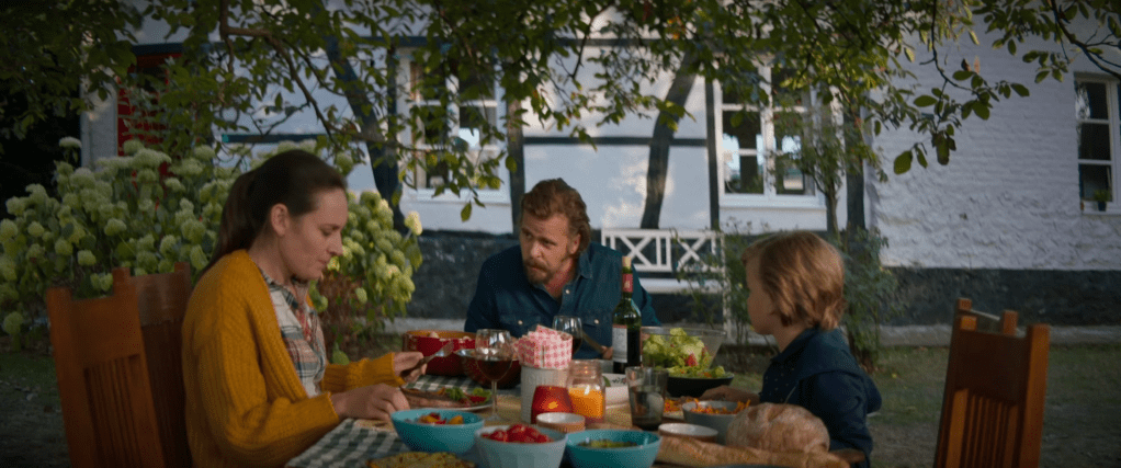 Still from Wild showing the family eating dinner outside in the garden of their rented countryside home. The woman is looking anxiously and nervously into the plate in front of her as the man is leaning and looking towards her. 