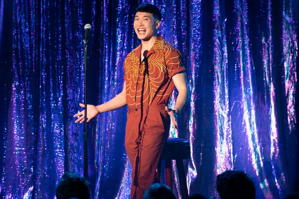 Still showing Joel Kim Booster on stage in the middle of a joke. With a blue/purple glittery backdrop, he is standing in front of his mic stand with a huge smile and his both arms outstretched to either side of his body.