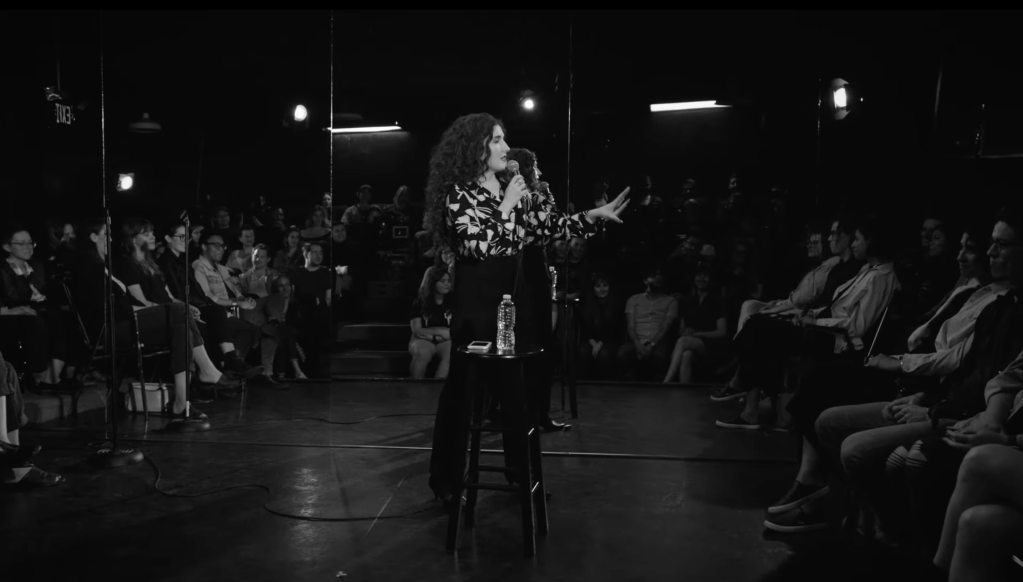 Black and white still from Cinnamon in the Wind showing performer Kate Berlant gesticulating in front of her crowd. The setting is intimate, as she is standing on the same level as her crowd, with mirrors acting as the backdrop and a small stool in front of her with phone and a bottle of water on top of it.