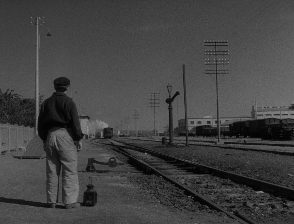 Still showing Guido from behind watching Moraldo depart from the seaside town. Down the railway tracks, Moraldo's train is visible with its smoke. Only Guido is there watching him leave. 