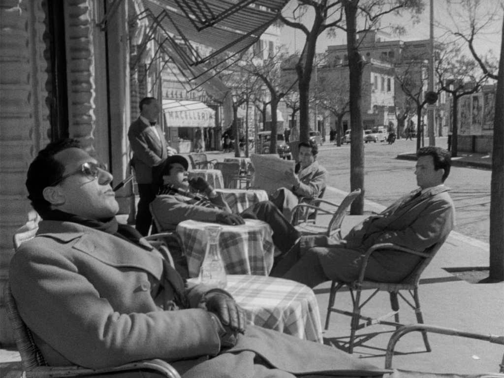 Still from I Vitelloni showing four of the five friends sitting outside of a café. One of them is reading a newspaper, one of them has his eyes closed as if he's sunbathing and the other two look deep in thought. They are together but feel separated. 