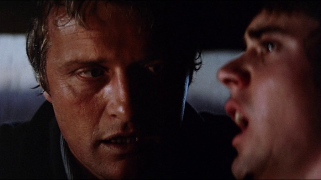 Close-up of John Ryder and Jim Halsey in the car during their first encounter. Ryder is in focus, hanging over Jim, clearly in control and power of the situation. Halsey is out of focus looking ahead in terror with his mouth open and eyes watered. 