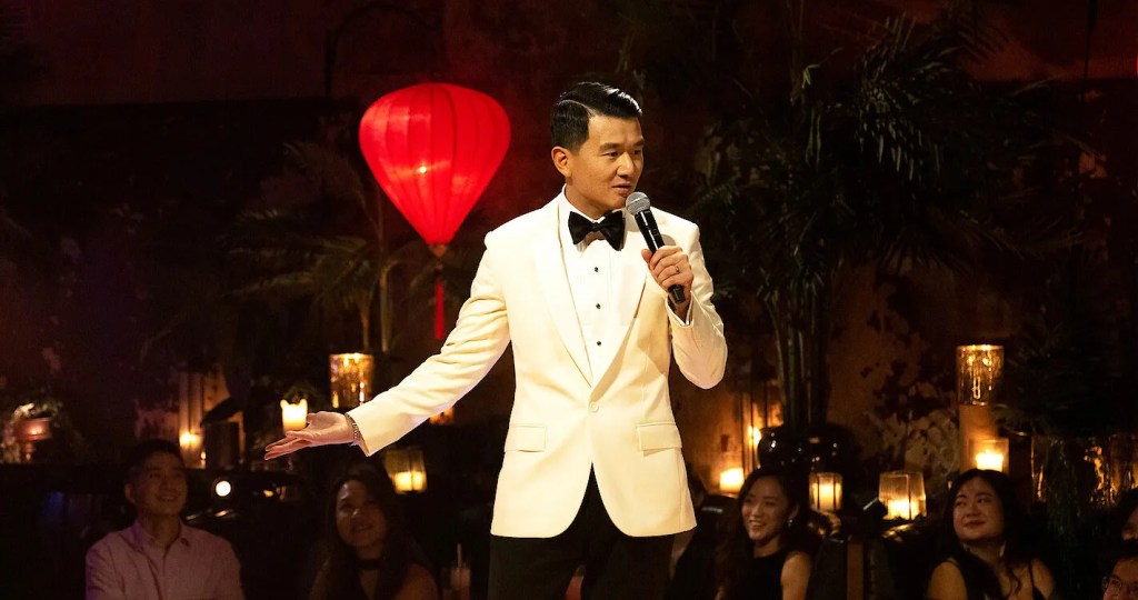 Still showing Chieng in the midst of telling a joke, as one hand is gesticulating and the other is holding the microphone close to his mouth. He is shown from the hip and up, showing his elegant white tux, black bow tie, and firmly slicked back and side parted black hair. Audience members are seen laughing in the background, and the setting is classy yet cozy, as he warm lighting and many candles makes it feel inviting. 