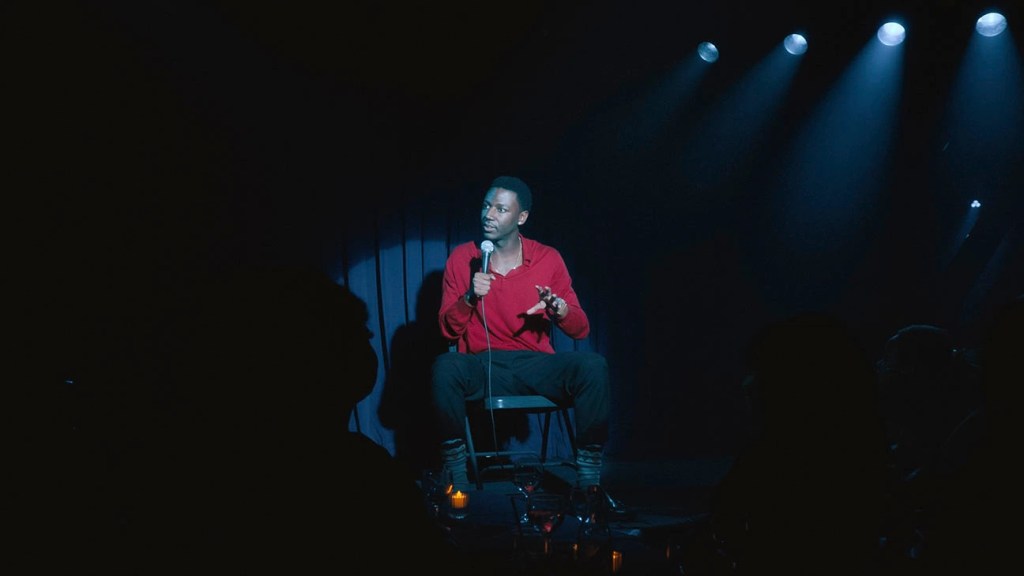 Still fully focusing on Carmichael as he sits on a folding chair and is caught in the midst of his act, gesticulating with one hand and holding his microphone with the other. Looking out towards the crowd, everything around him is out of focus and in darkness, as the spotlights hits him and puts him at the centre. He is wearing a red jumper, black pants, patterned socks and black loafers.