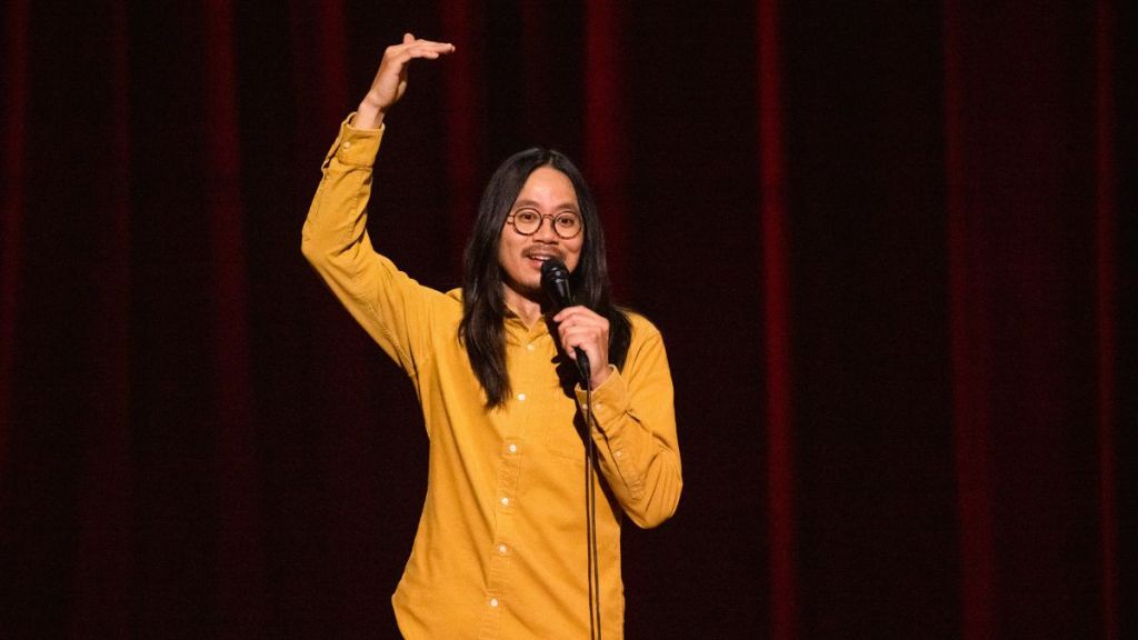 Still showing Wang wearing a bright yellow shirt with white buttons, his long dark brown hair out and round glasses. He is shown from the hips and up, with a dark red backdrop. He is shown caught in the middle of a joke, holding the microphone with one hand with the other hand raised above his head as he is trying to illustrate something. He has a happy facial expression.