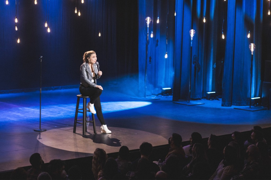 Still from Look at You taken with some distance to its performer Taylor, as she sits on a stool on a stage looking upwards whilst speaking into her microphone. The stage features decorative lightbulbs hanging from the ceiling as well as spotlights — one of them directed directly at Taylor — as sources of light. On the bottom of the still, audience members are seen looking up at Taylor.