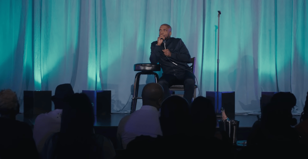Still showing Siddiq sitting on a chair on a stage in front of his audience, with an empty mic stand to the right whilst leaning his arm and head on a stool next to him to the still's left. He looks deep in thought, contemplative, with one hand leaning on his chin and mouth and the other holding the microphone.