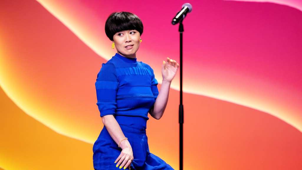 Still showing Okatsuka from the hips and up, as she's bending her knees and pushing her butt out, as if caught doing a dance. She is wearing all blue and bright yellow earrings and painted nails. Out of focus in front of her is her mic stand with her microphone. Behind her is a neon backdrop consisting of three waves in yellow, orange, and pink.