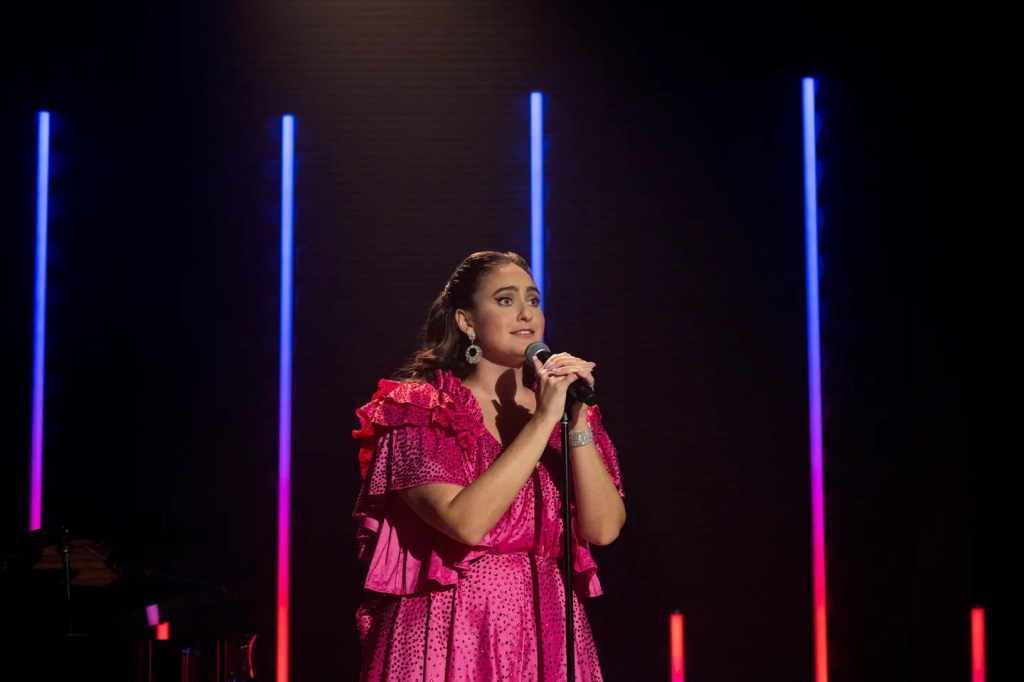 Still showing Catherine in her pink rhinestone romper with an excited facial expression as she is holding both of her hands on the microphone in her mic stand, reading to start performing her song at any second. There's hope in her eyes, as she looks out towards the crowd in front of her that's outside of the frame. 