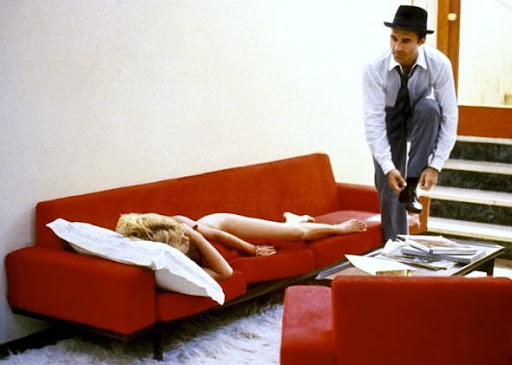 A still from the film 'Contempt' depicting a woman lounging in the nude on a red sofa, and a fully dressed man appearing to be in the act of taking off his shoes and observing her.