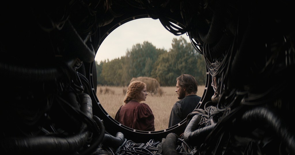 Maura and Eyk climbing into a memory. They are positioned within a circle as the shot is taken from inside the cylindrical pathway they have just exited. Within the pathway, visible cables of various sizes are seen. Maura and Eyk are looking at each other with serious facial expressions. In front of them is a hay field with lightly browning grass as well as a green forest in the distance and a bright, almost white, coloured sky.