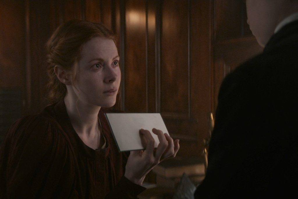 Maura, with tears in her eyes, holding up an envelope to Elliot (also referred to "The Boy") who is outside of the frame, only seen partially. Maura's red hair is pushed back in a braid and she is wearing a maroon dress. They are within one of the cabins.