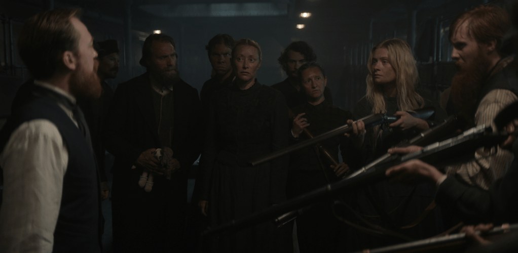 Mutiny erupts onboard the ship. Here third-class passengers are shown in control, holding guns and directing them as well as their stern facial expression towards the ship's crew that is standing to the left in the frame. The only one of the third-class passengers that look concerned is Anker, who is standing somewhat in the middle looking not at the crew but at his wife and daughter instead.