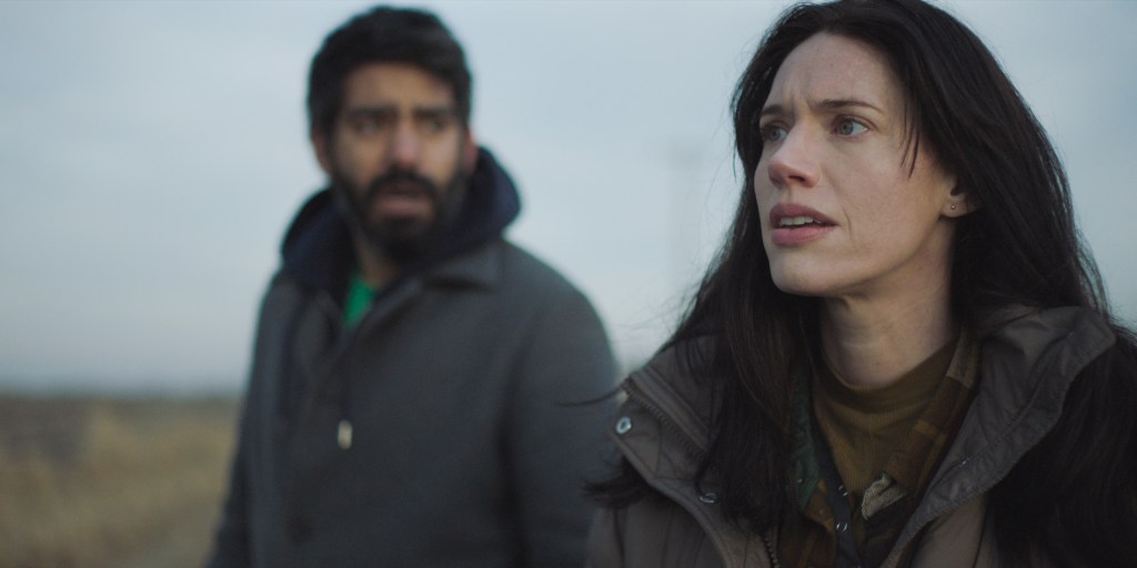 A still of Rose (Katie Parker) in 'Next Exit'. She stands on an open road with blue sky behind her, and Teddy (Rahul Kohli) is out of focus in the background.