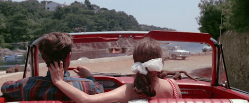 A still from 'Pierrot le Fou' depicting a man and woman seen from behind in a red car. The woman puts her left hand on the man's neck as they drive down a road.