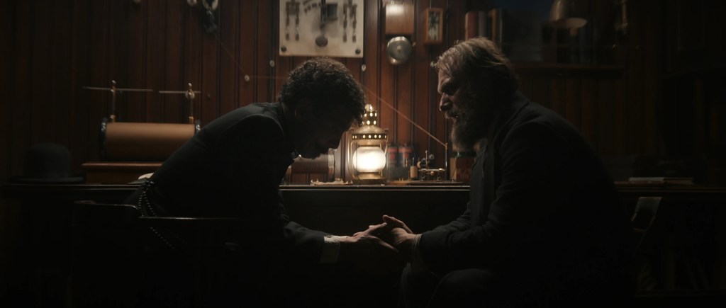 Ramiro and Anker sitting down, facing each other. Ramiro is holding Anker's hands in his, looking down at them in a moment of silence. Anker, with a more distressed facial expression, is looking at Ramiro as if awaiting his response. Behind them, centered between them, is a lit lantern which barely lighten up their faces.