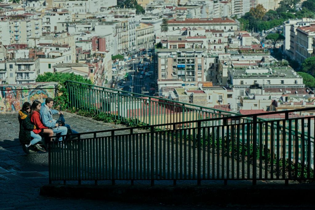 Still showing the differences between neighbourhoods in Naples. Giovanna is seen riding with two friends on a vespa. In the background buildings, roads and big apartment complexes are seen. The still is taken from a distance and from above where Giovanna and her friends begin their ride down to the more levelled roads. It puts a perspective on these three young characters and much more there is to discover beyond their own worlds.