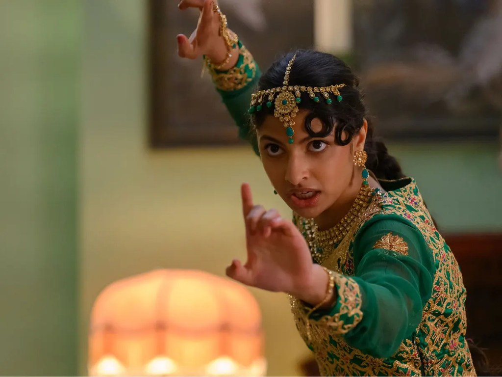 An Indian woman from Polite Society in traditional dress, green, with a jewelled headdress, arms raised in a fighting pose and a serious expression on her face