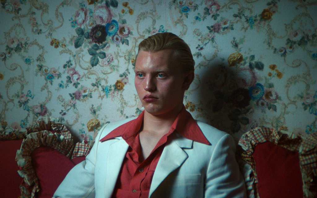 Nicklas (Andreas Lykke Jørgensen) seen sitting on a couch in a medium close up shot. His facial expression is firm, one can see that he is clenching his jaw and focusing hard on something as his veins in his forehead are emphasised. His blonde hair is slicked back. He is wearing a red button-up shirt and a white blazer. He is facing and looking at something out of frame. Behind him and the couch is a wallpaper filled with flowers.