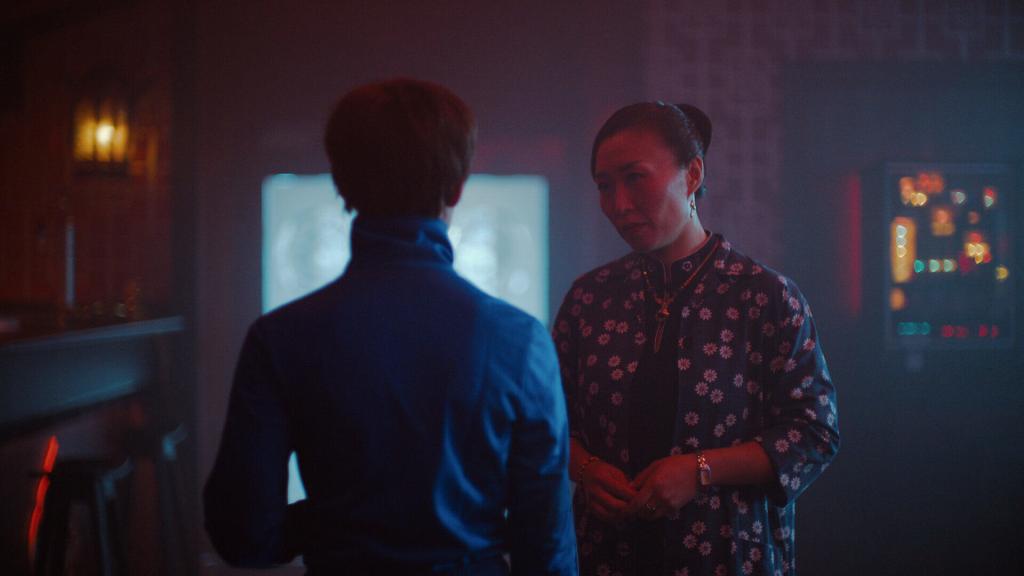 Miu (Angela Bundalovic) and Mor Hulda (Li Ii Zhang) standing in her restaurant. Miu has her back facing the camera, whilst Mor Hulda is seen standing slightly in front of Miu, facing her, and looking at her. Her hands are joined together at the her stomach. 