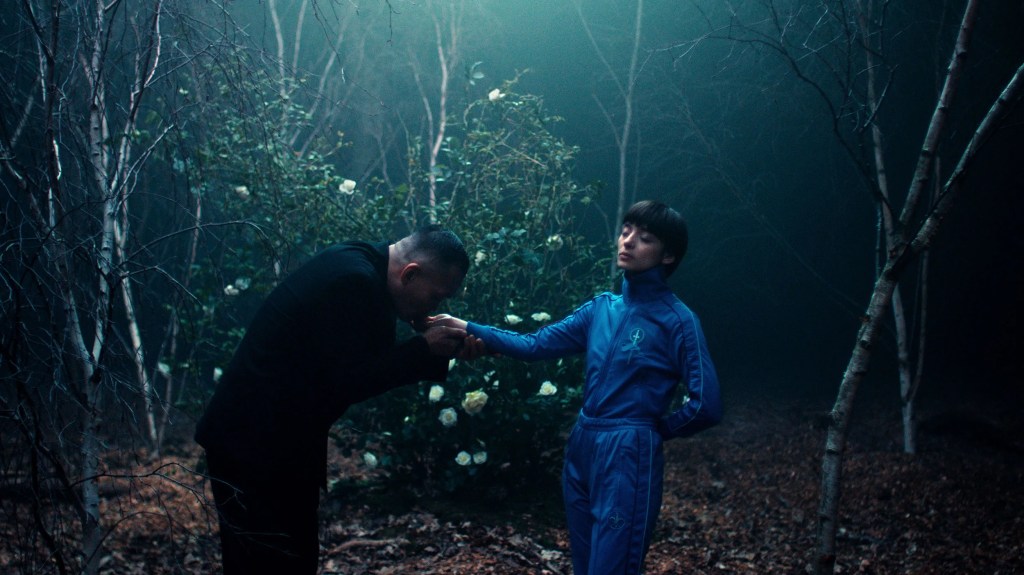 Miu and Chiang (Jason Hendil-Forssell) seen standing in a forest. Chiang is bowing to Miu as he is holding her right hand in his and lightly kisses it. He is wearing all black. Miu is wearing her blue tracksuit. Her eyes are closed and her head is slightly tilted back as her left arm is behind her back. A bush with white roses is seen behind them. 