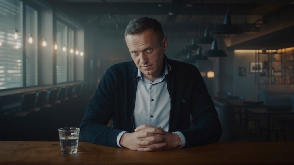 Still from "Navalny" showing Alexei Navalny sitting down in front of the camera with a glass of water next to him, as he is looking directly into the camera. His hands are locked together in front of him, whilst leaning on the table. 