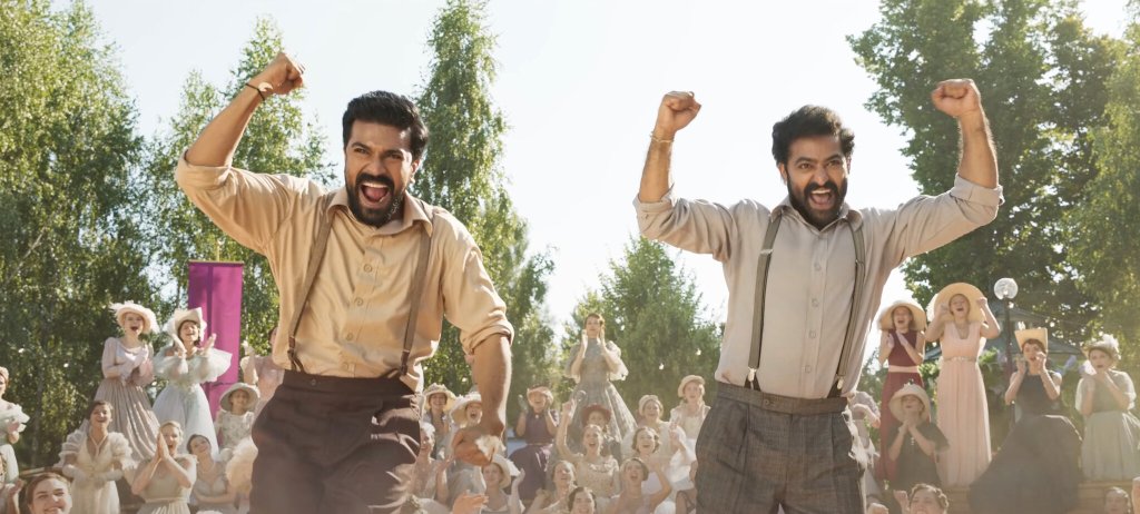 Still from "RRR" showing its two main character engaging in the big musical number featuring the hugely successful song "Naatu Naatu." They are both very excited and happy, raising their fists above their hands, as they are caught in the midst of dancing. In the background, various women are seen in upper-class clothing cheering them on. 