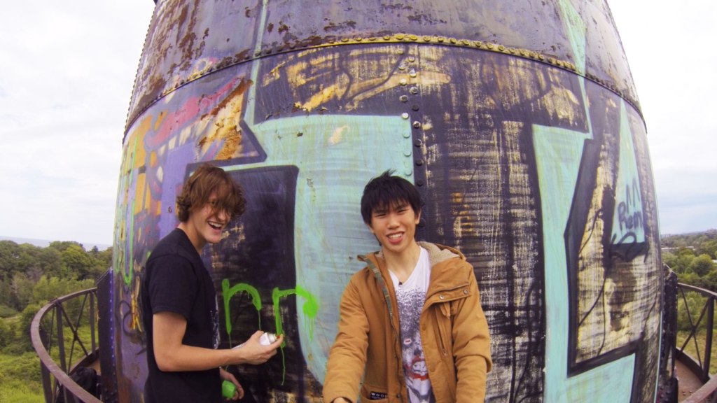Still showing Justin and Ethan standing on the landing platform of a water tower, which is a circular building. They are both smiling, happy and mischievous as Justin is holding a can of lime green spray paint, fully caught in the act of writing something on the property (which is already filled with half-faded graffiti from previous visitors). Ethan is the one filming, using an accessory of some kind (like a selfie stick) to get a wider shot of the two, as they both look directly into the camera. They are up several meters in the air.
