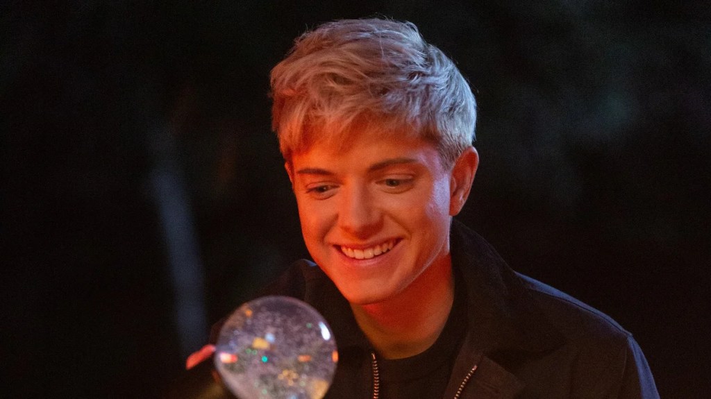 Close up of Mae Martin smiling big with teeth as they are looking at a snow globe they are holding up in front of their face. They are sitting outside, wearing a jacket, and there's a warm light lingering on Martin's face from the campfire in front of them (that is outside of the frame).