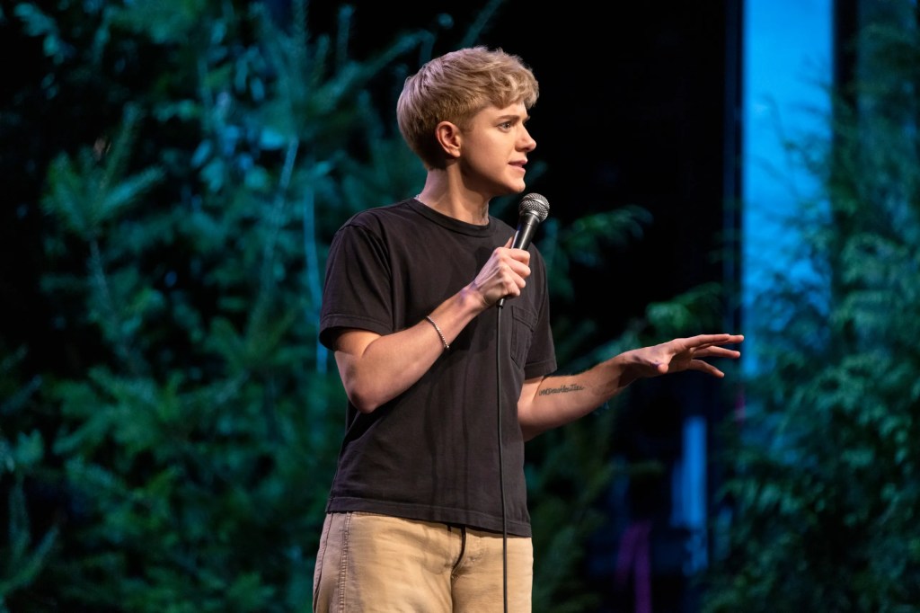 Mae Martin standing on stage performing 'Sap.' A medium close up showing their face in profile. They are in the midst of talking about something, with a focused facial expression and one arm stretched out in front of them whilst the other is holding the microphone close to their mouth. They are wearing a black t-shirt and yellow/beige pants. Behind them, trees and greenery are shown as part of the stage.