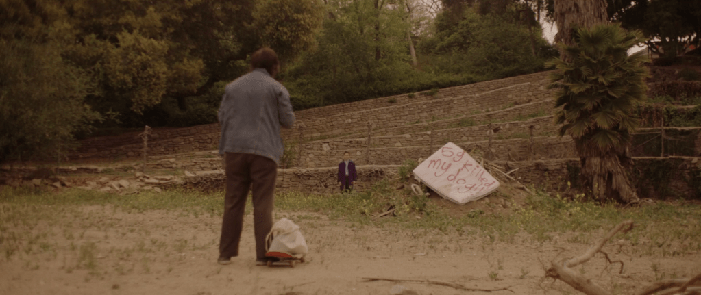 Tatum as Clay shown from behind photographing. Beside him on the ground is his skateboard with his tote bag on top of it. In the distance, Thomas and Whit is seen wearing his purple trench coat. Next to him is an abandoned mattress with the words "5g killed my dog" spray painted on top of it. No other people are around, they are in a quite rural-looking place, with trees in the background as well as rows of stones. 