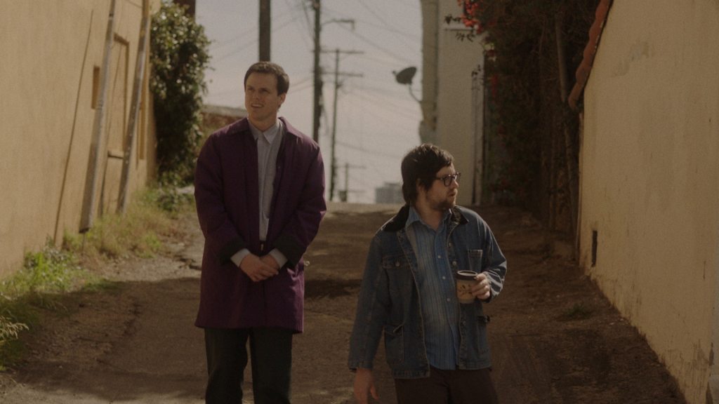 Whitmer Thomas (left) and Clay Tatum (right) walking down a street in LA. The light is warm and yellow. They are facing the camera, but looking to either side of themselves. Tatum is shorter than Thomas. Tatum is wearing a denim jacket and Thomas is wearing a dark purple trench coat. Tatum is holding a to-go coffee mug in one of his hands.