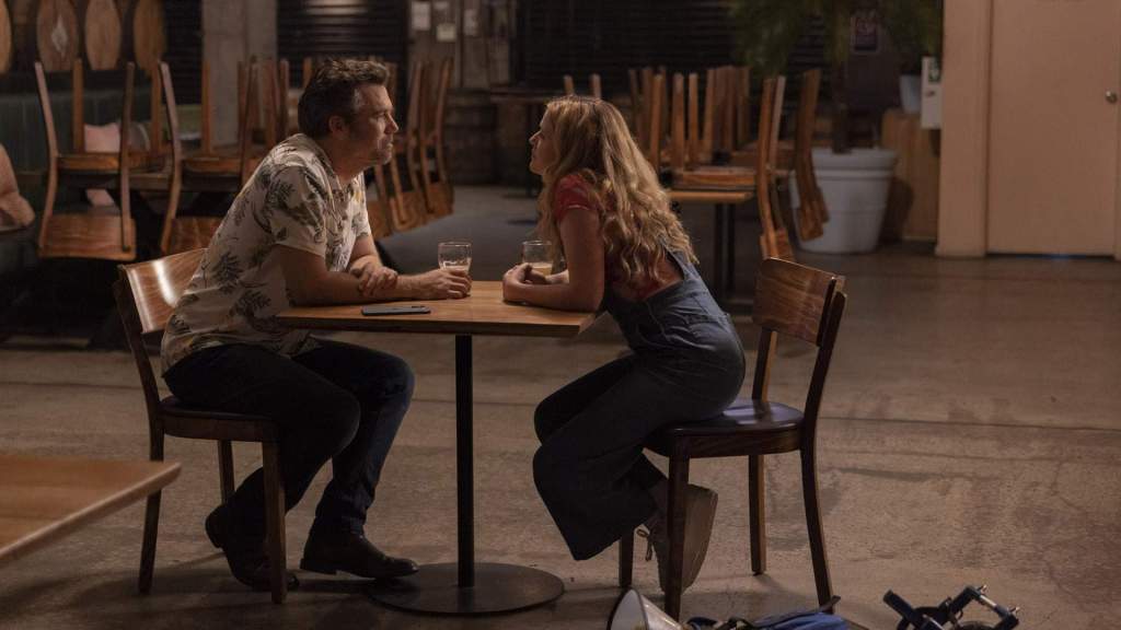 Still showing Ashley and Gordon sitting on either side of a table, facing each other and smiling towards each other. The interior is from Gordon's brewery, but it's off hours and closed, so Ashley and Gordon are alone and the light is slightly dimmed. Behind them are other tables shown with their respective chairs put on top of them. They are drinking beer and the dog is just outside of the frame. 