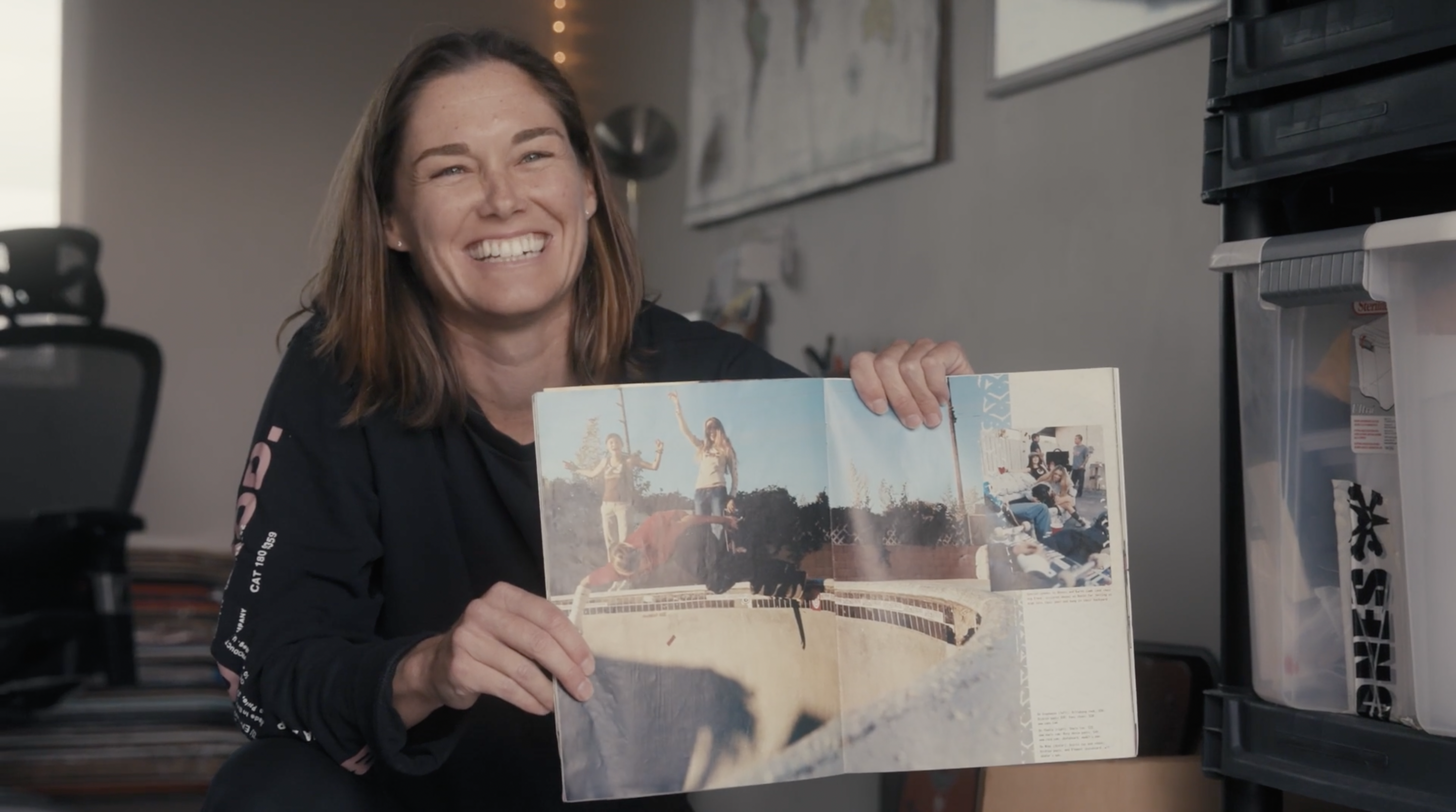 Mimi Knoop smiling big with visible teeth and looking towards the camera whilst holding up a skate magazine, showing a spread where a photograph of her was covered. She is seen skating in a swimming pool as two models stand on the edge cheering with their hands in the air.