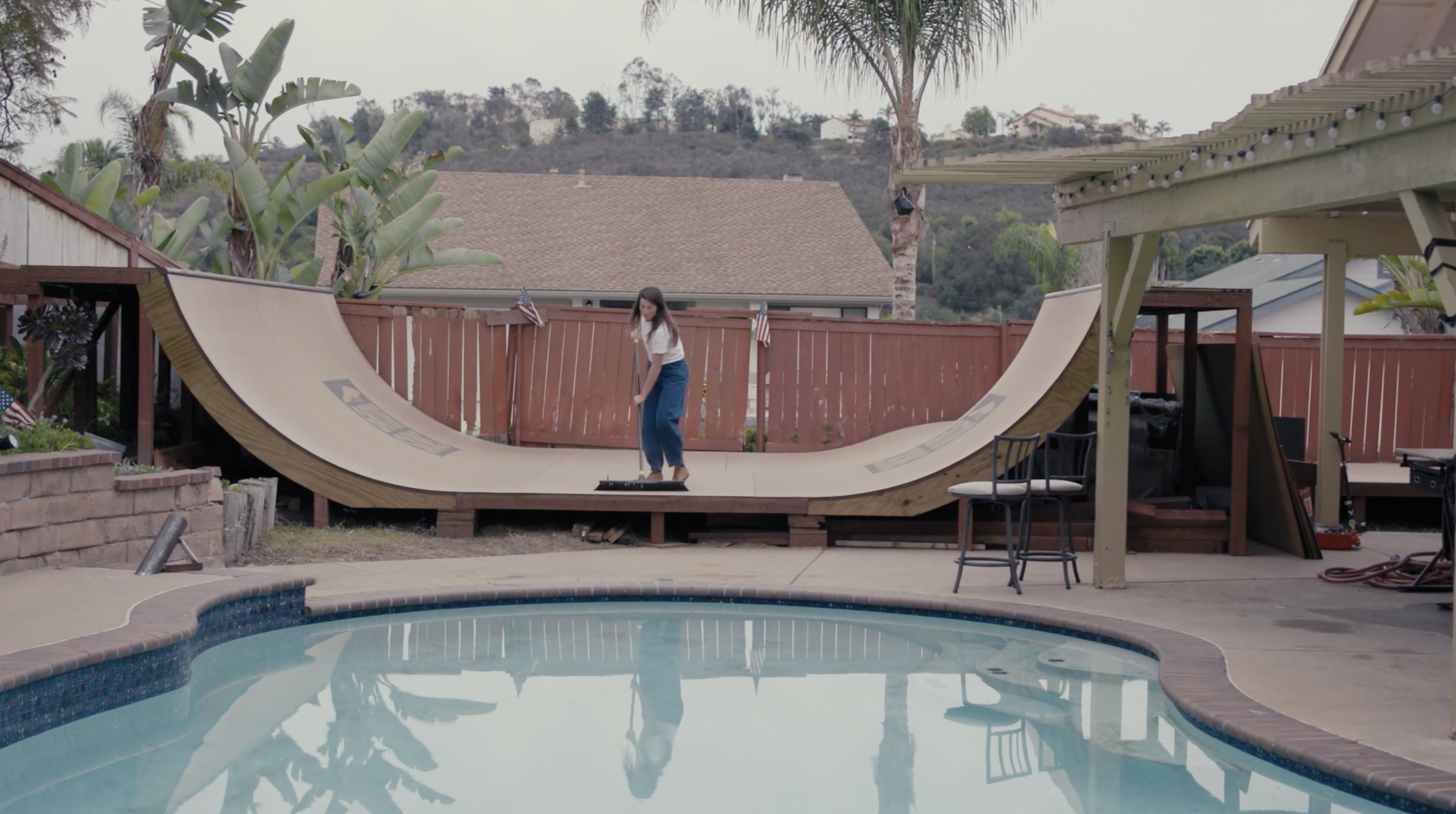 Nicole Hause at her and Nora's house at the time of filming. She is in the backyard, cleaning their skate ramp with a wide sweeper. Her body is facing the camera, as she looks down, concentrated on her work. Ahead of her are the curves of a filled swimming pool seen.
