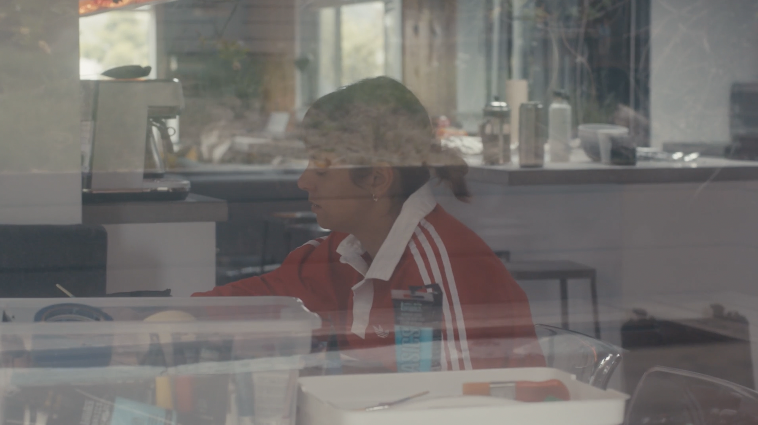 Nora Vasconcellos captured from outside (a glass window separating her from the camera) as she is sitting inside painting. She is wearing a red Adidas shirt with a white collar and white details. Next to her a boxes of art supplies seen, including various tubes of paint and brushes. It feels as if she is caught in a moment of positive solitude and quiet.