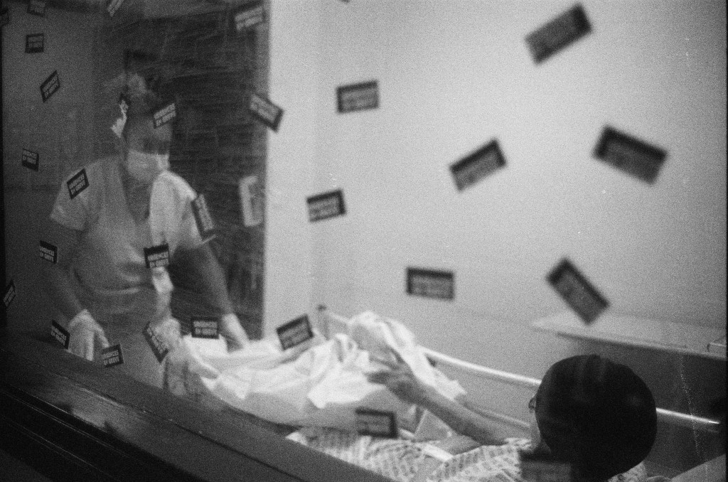 Black and white still. The image is captured from one side of a window, as a medical worker and patient (seen lying in a bed) are on the other side of the window. The window is covered with stickers with hard-to-read French text (possibly saying "urgences en grève" which translates to "emergencies on strike").