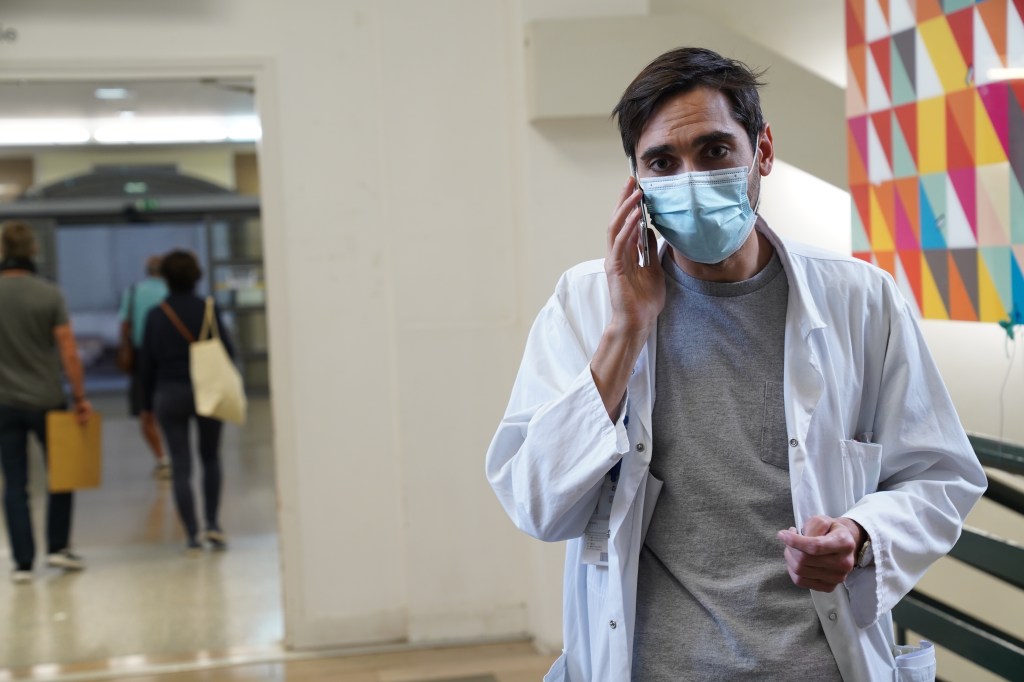 Still in colour showing Jamal from the waist and up inside one of the corridors of the Beaujon. He is wearing his white coat with a grey T-shirt underneath it. He is wearing a medical mask and is talking on his phone. His facial is slightly concerned, as he looks disheartening towards something in front of him whilst slightly frowning. He is the one in focus.