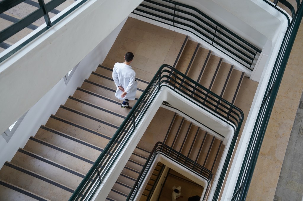 Shot from inside the Beaujon hospital, as Jamal with his white doctor's coat walks by himself down a set of stairs. The still is taken from above, underlining how many floors the hospital has and how Jamal walks up and down them all day long.