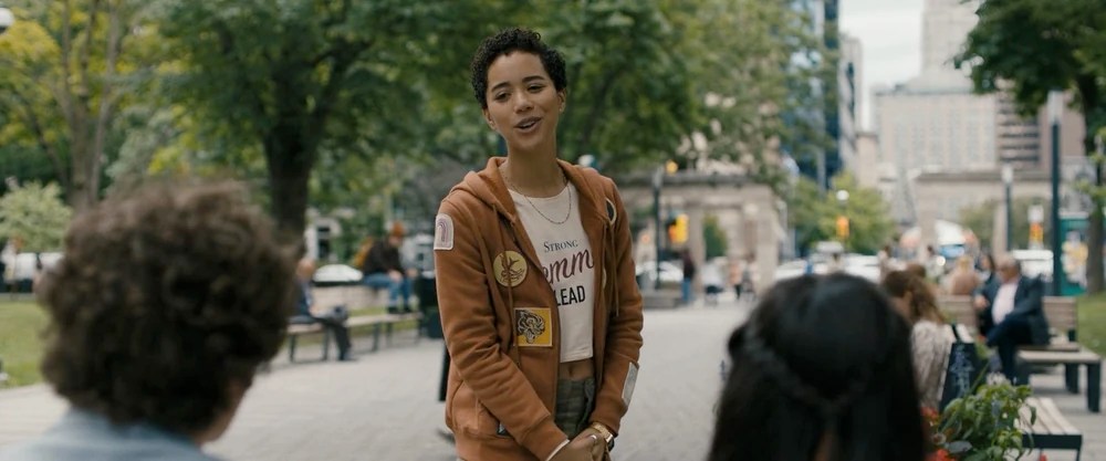 A still of (Jasmin Savoy Brown) in 'Scream VI'. She is outside wearing a jacket adorned with various patches, standing up as she addresses two other characters who are sitting down seen from behind.