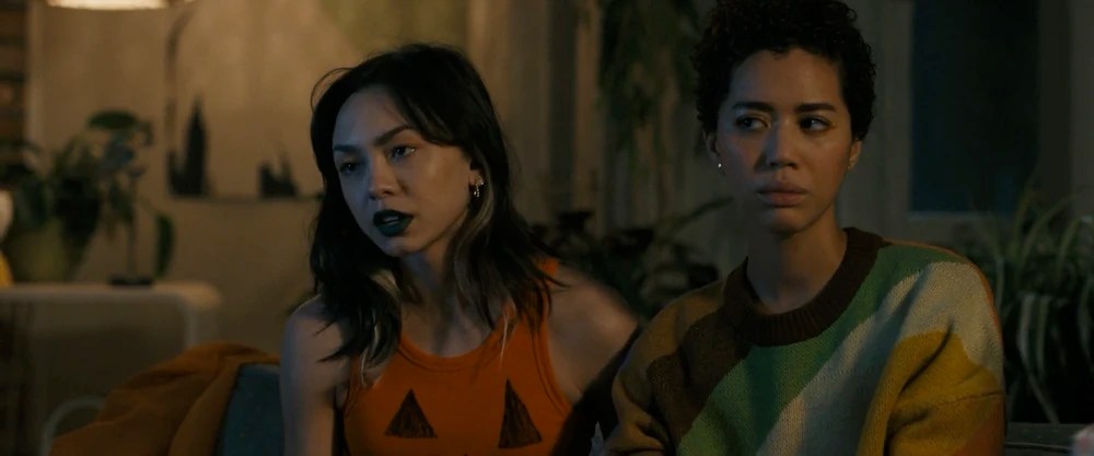 A still from 'Scream VI' of Anika (Devyn Nekoda) and Mindy (Jasmin Savoy Brown) sitting on a couch. Anika wears an orange tank top with a jack o'lantern face, while Mindy wears a colourful oversized sweater.