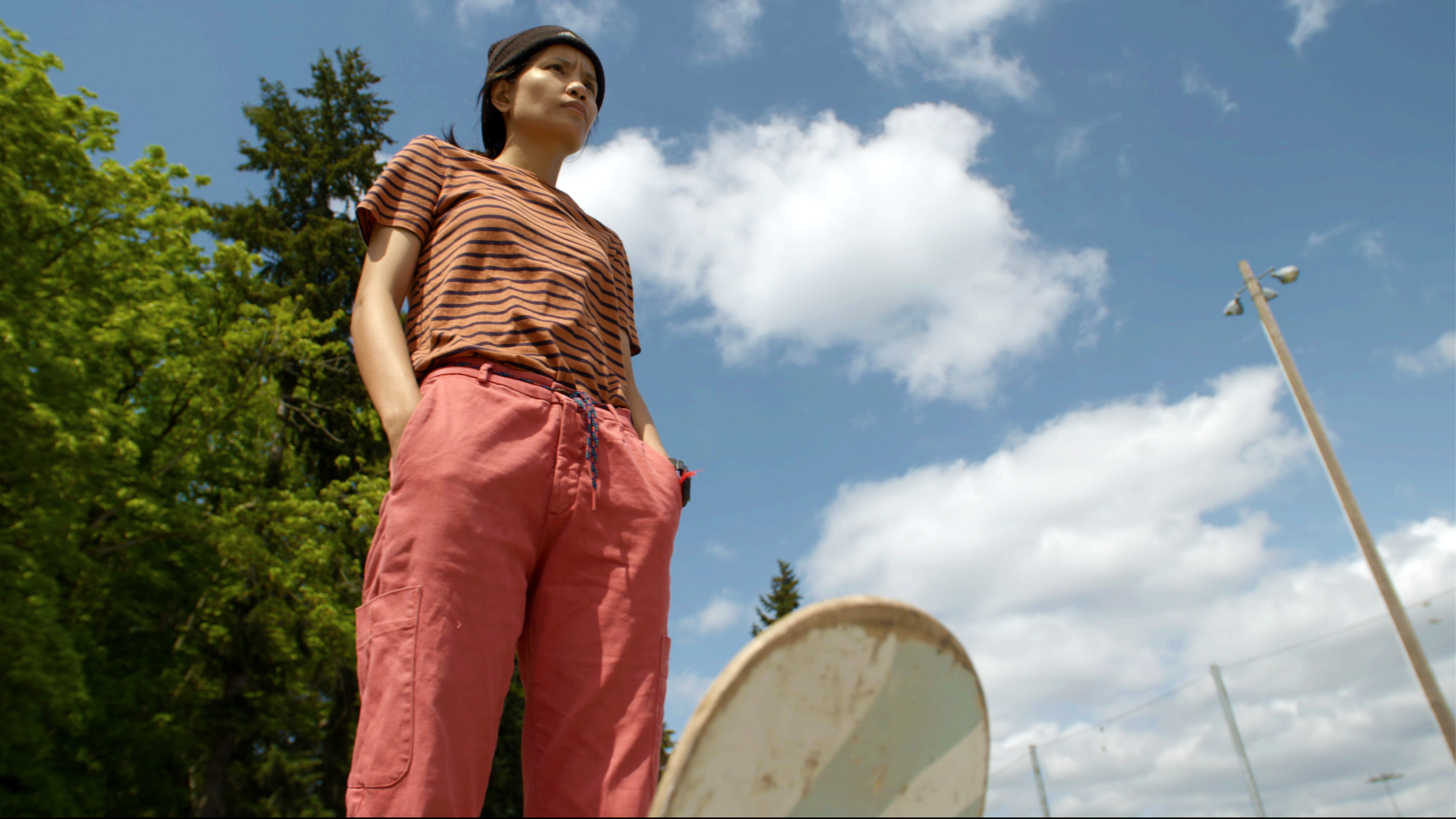 Tin shown from below, a low angle of the camera, as she is looking with a concentrated facial expression ahead of her, as if deciding where to skate next. She is wearing a dark beanie, a striped t-shirt and baggy cargo pants. The top of her skateboard deck is shown in the frame. Behind her is a blue sky with white clouds and greenery present.