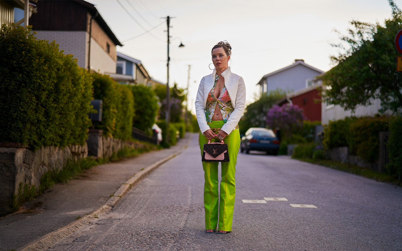 Amelia is seen standing left all alone on a street. She is wearing Y2K fashion, including sandals, lime green pants with a low waist, a top that shows off her belly piercing and over it a white short coat. It is an outfit that stands out within the frame. She is looking sad and distressed. 
