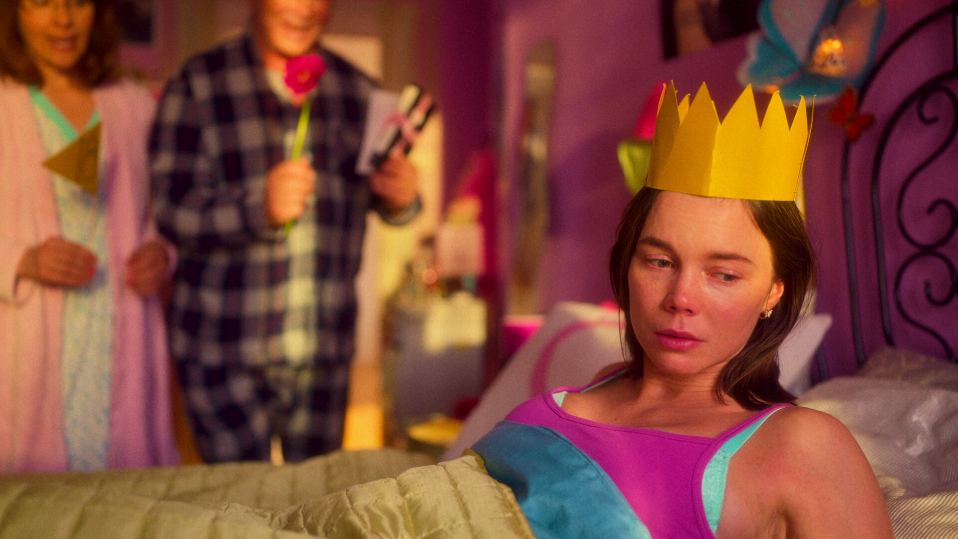 Amelia shown waking up in her childhood bedroom on her eighteenth birthday. She is wearing a gold-coloured crown made in paper and looks distressed as she turns her head away from her parents. Her parents are standing next to her bed, out of focus, in the midst of singing and celebrating Amelia. The father is holding a flower, an envelope and a VHS cassette and the mother is holding up a little flag that says "18".